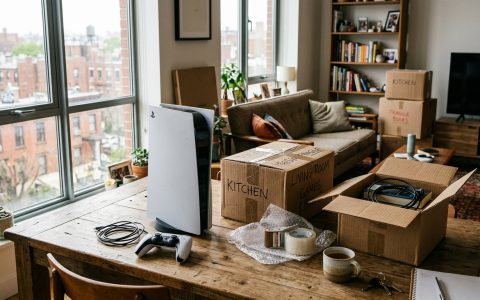 A PlayStation 5 console and controller next to unpacked moving boxes in a second-floor apartment