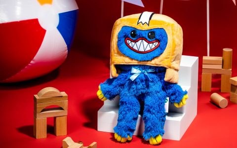 A blue Huggy Wuggy plush wearing an orange Boxy box head, sitting on a white step among wooden blocks and a beach ball