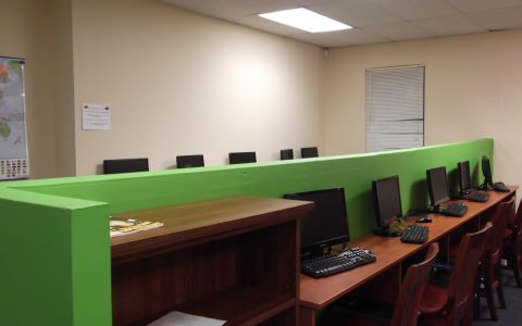 An empty computer lab with rows of monitors and keyboards on wooden desks behind a green partition
