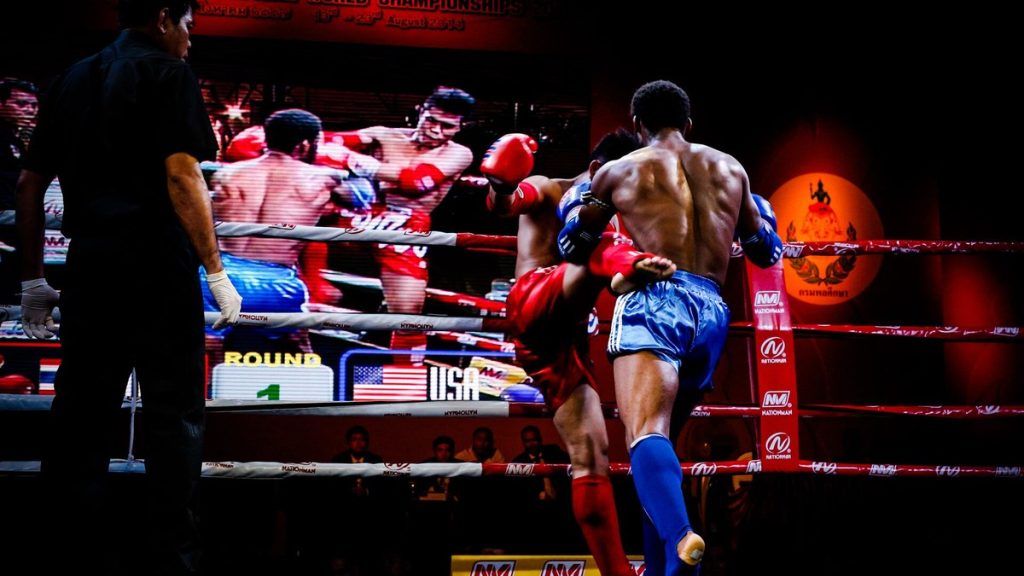 Two Muay Thai fighters exchange kicks in a ring as a referee watches