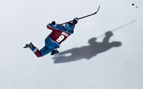 Overhead view of Cale Makar in a Colorado Avalanche jersey taking a shot on white ice