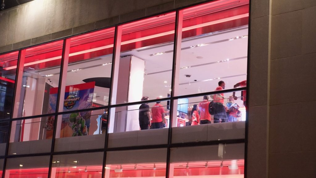 Staff and shoppers inside the Nintendo New York store, seen through its red-lit windows at night