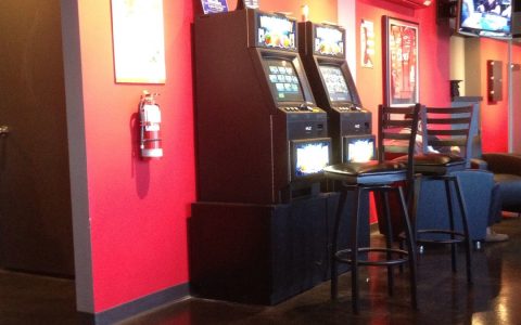 Two video gaming terminals against a red wall in a bar, with bar stools in front